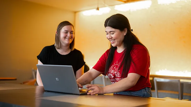 Two female students smile, with one working on a laptop