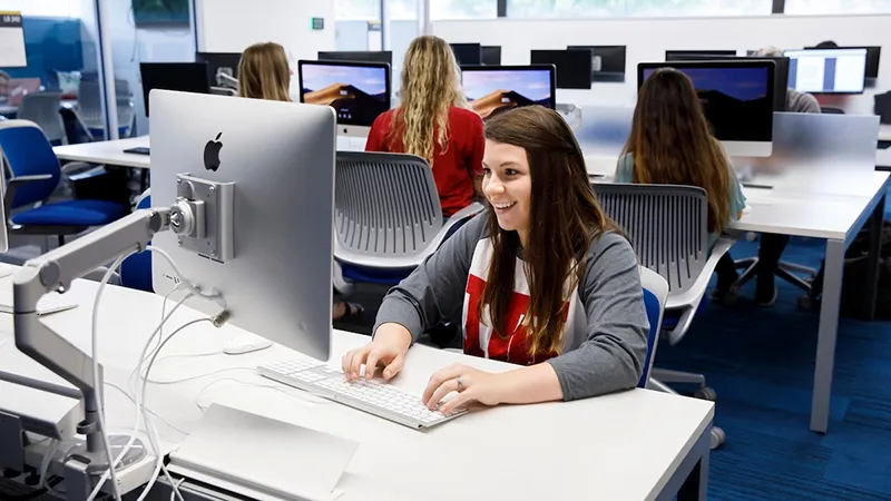 Student smiles while using a desktop computer in a busy computer lab with other students.