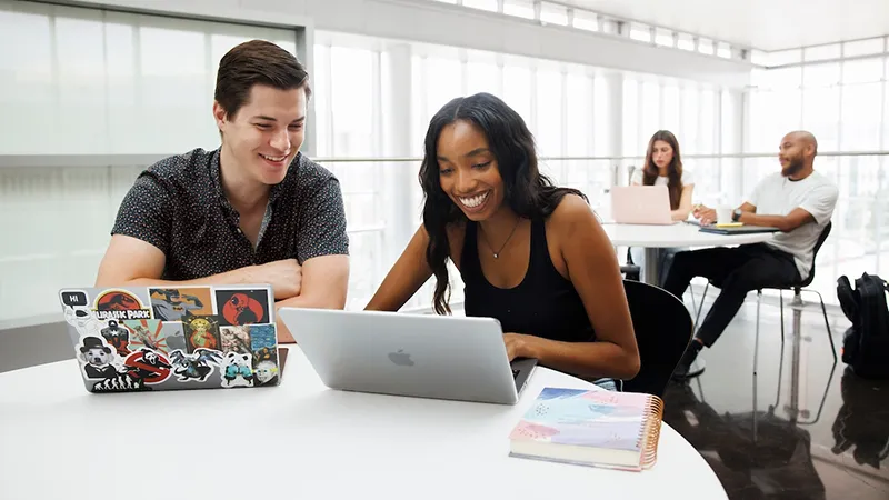 Four students study in a common area on laptops, smiling and working in pairs.