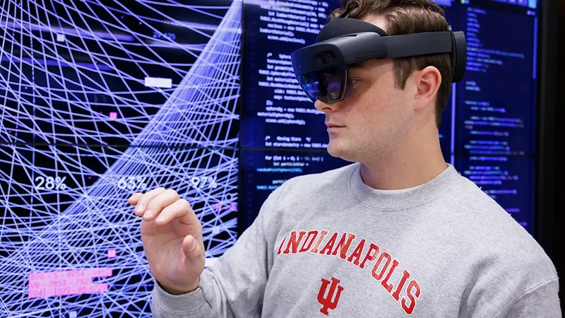 A male in an IU Indianapolis sweatshirt wears VR glasses in front of a media wall with code and graphics.