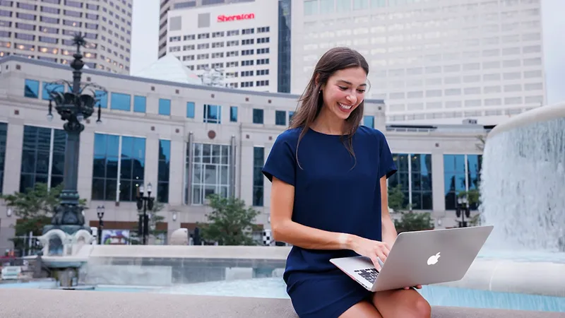 A female works on a laptop outside, sitting by fountains with a city district background.